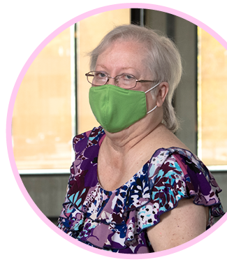 Barbara standing along the railing outside the the Cancer Resource Center, she is wearing a green face mask and a purple floral shirt. 